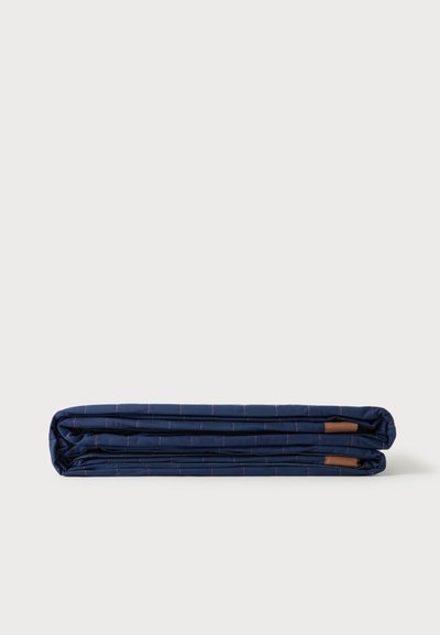 Couette bleu marine soigneusement pliée avec de fines lignes de quadrillage roses et de petites pièces en cuir marron sur fond blanc.
