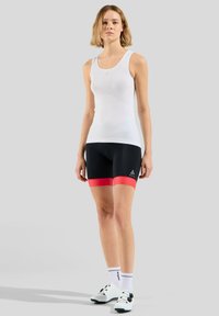 Witte mouwloze tanktop met mesh-textuur, gecombineerd met zwarte aansluitende shorts met rode accenten. Model draagt fietsschoenen en sokken.