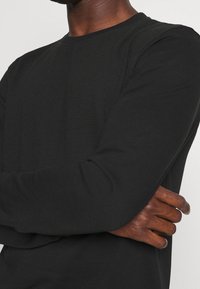 Svart långärmad skjorta med ett strukturerat mönster på framsidan, rund halsringning och passformade muddar. Slät tyg med en diskret lyster.