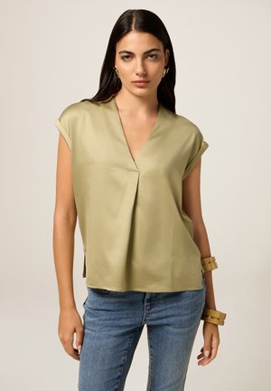 Donna con lunghi capelli scuri che indossa una blusa corta con scollatura a V di un leggero olivastro, jeans blu, orecchini dorati e braccialetti abbinati sul polso destro.