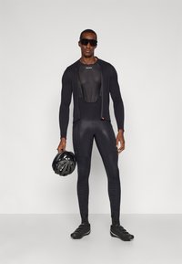 Costum de ciclism negru, cu un top din material texturat, ajustat, cu mânecă lungă și colanți tip bib. Modelul ține în mână o cască neagră și poartă ochelari de soare întunecați.