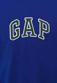 Mėlynas medvilninis marškinėlis su žodžiu "GAP" baltomis ir tamsiai žaliomis siuvinėtomis raidėmis per krūtinę.