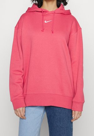 Osoba ubrana w za dużą różową bluzę Nike z kapturem i ściągaczami oraz dżinsy w dwóch odcieniach niebieskiego na jednolitym tle.