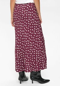 Gonna maxi bordeaux con pois bianchi, realizzata in tessuto morbido, presenta un design fluido e una texture liscia. Indossata con stivali neri alla caviglia.