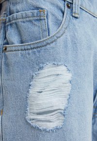 Jeans de mezclilla azul claro con un gran parche desgastado y bordes deshilachados. Costuras expuestas y un clásico diseño de cinco bolsillos.