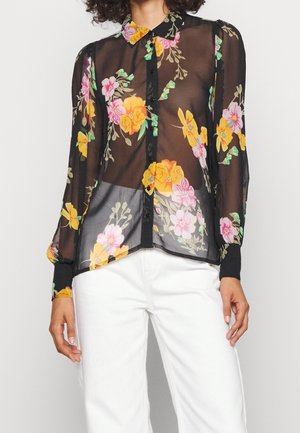 Zwarte doorschijnende blouse met lange mouwen en een bloemenpatroon in geel, roze en groen, gedragen met witte broek.