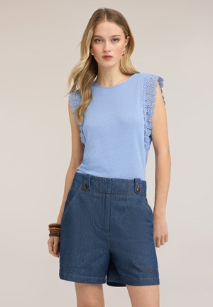 Mujer que lleva una camiseta sin mangas azul claro con ribete de encaje combinada con pantalones cortos de mezclilla oscuros de talle alto, de pie sobre un fondo neutro.