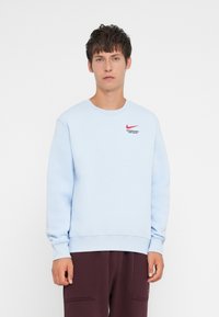 Light blue sweatshirt featuring a crew neckline and cuffs, with a red Nike logo and text on the left chest. Fabric has a soft texture.