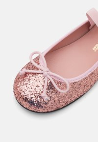 Bailarina rosa con brillantina, con punta redonda, cinta elástica y detalle de lazo. El zapato tiene un interior acolchado y una suela de caucho negra.