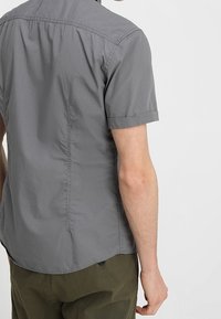Grijze shirt met korte mouwen van lichtgewicht stof. Heeft een kraag, een iets afgeronde onderkant en zichtbare stiksels langs de rug- en zijnaad.