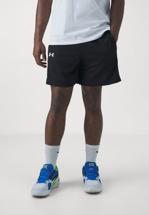 Homme portant un short noir Under Armour, des chaussettes blanches et des baskets Puma bleues, blanches et vertes, debout sur un fond clair uni.