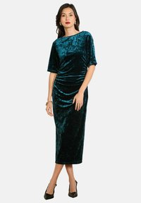 Tyrkysové sametové midi šaty s krátkymi rukávmi, so zbieraním v páse a hladkým povrchom. Skombinované s čiernymi lodičkami s ostrou špičkou.