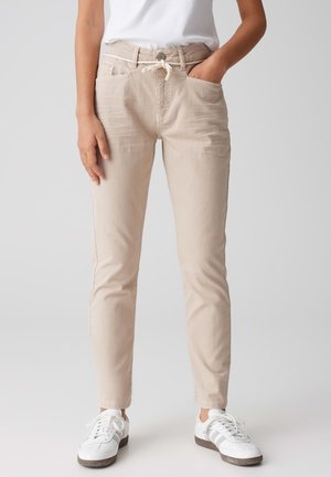 Beige Baumwollhosen mit einem Kordelzugbund, zwei Vorderaschen und einem schmalen Schnitt, kombiniert mit weißen Sneakern vor einem neutralen Hintergrund.