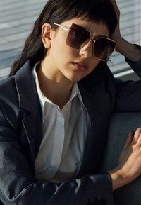 Ochelari de soare pătrați cu lentile întunecate, accente aurii și un cadru subțire, purtați de o persoană îmbrăcată într-un sacou gri și o cămașă albă.