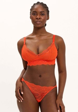 Vrouw die een feloranje kanten bralette en bijpassend kanten bikiniondergoed draagt, staand tegen een effen lichte achtergrond.