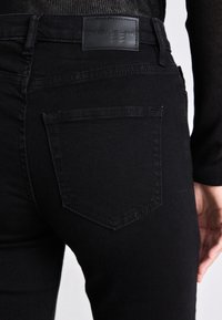 Sorte denimjeans med synlig baklommestikking og et lærmerke merket "DKNY JEANS" båret av en person.