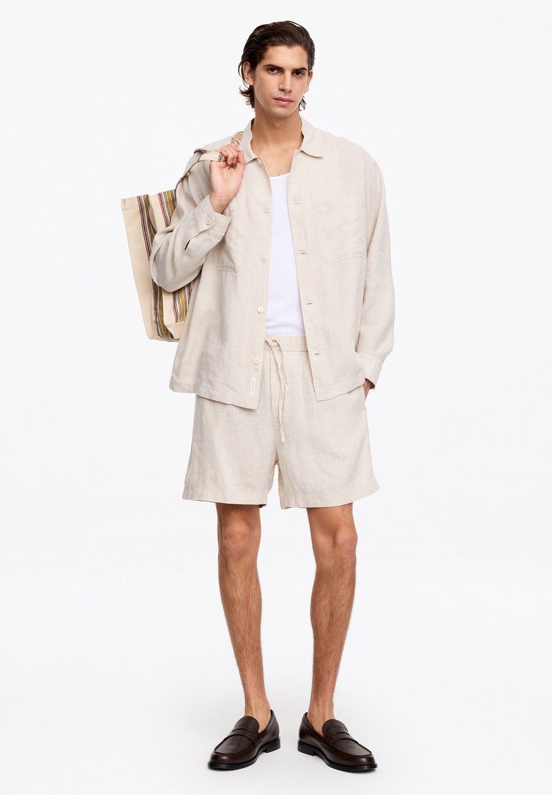 Homme portant une chemise beige à boutons et un short, un débardeur blanc, des mocassins foncés, tenant un sac cabas rayé sur l'épaule, debout sur fond blanc.