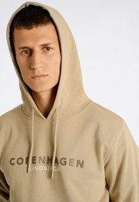 Felpa beige con tasca a marsupio frontale, cordino attorno al cappuccio e scritta stampata "Copenhagen Lindbergh" in marrone scuro e bianco.