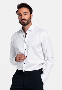 Weißes Langarmhemd mit taillierter Passform, blauen Knopfakzenten und einem klaren Kragen. Glattes Material mit einem dezenten Glanz, kombiniert mit dunklen Hosen.