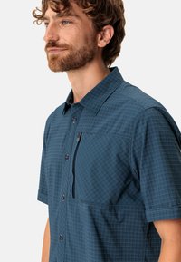 Camisa de manga corta en tela azul oscuro con un patrón de cuadrícula, que cuenta con un cierre frontal de botones y un bolsillo en el pecho con cremallera.