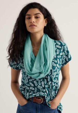 Jauna sieviete ar gariem cirtainiem matiem, tērpusies zili zaļā rakstainā īsroku krekliņā, gaiši zilā šallē, brūnā jostā un zilās džinsu biksēs, ar rokām kabatās.