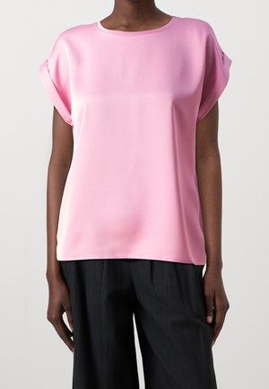 Kobieta ubrana w luźną, krótką, różową bluzkę wpuszczoną na spodnie, stojąca na jednolitym tle, w czarnych szerokich spodniach.