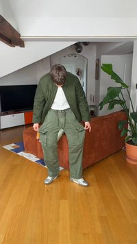 Persoană îmbrăcată în geacă verde și pantaloni cargo, privind în jos într-un living cu podea din lemn, canapea portocalie, televizor și o plantă mare în ghiveci.