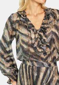 Blusa transparente en tonos apagados de marrón, gris y negro, con escote en V, borde rizado y botones decorativos. Mangas largas con puños fruncidos.