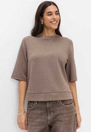 Kvinde med langt mørkt hår iført en taupe kortærmet top, brune jeans, et sølvhalskæde og sølv øreringe, smilende og kigger til højre.