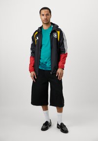 Mand står iført en sort, rød og gul sportsjakke, teal skjorte, sort brede shorts, hvide sokker og sorte sko på hvid baggrund.