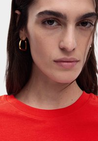 Rood T-shirt met korte mouwen en een ronde hals, met een gladde textuur en contrasterende huidskleuren. Gouden creolen accentueren de look.