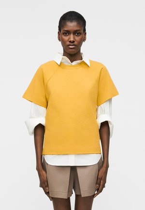 Γυναίκα όρθια, στραμμένη προς τα εμπρός, φοράει κίτρινο τοπ με κοντά μανίκια πάνω από λευκό πουκάμισο με γυρισμένα μανίκια και μπεζ σορτς.