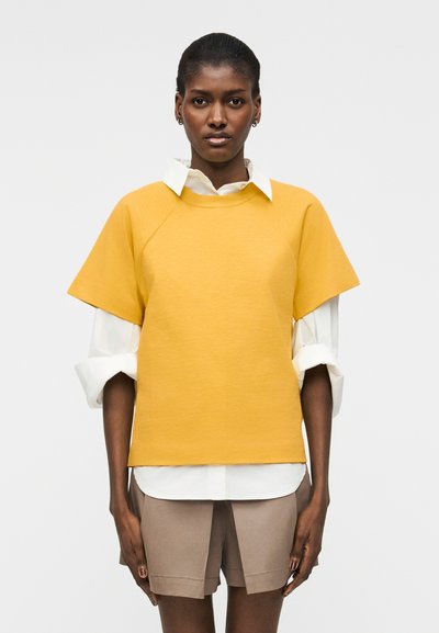 Femme debout, face à l'avant, portant un haut jaune à manches courtes superposé à une chemise blanche aux manches retroussées et un short beige.