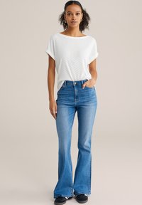 Joven con cabello rizado, vestida con una camiseta blanca de manga corta, vaqueros azules acampanados de talle alto y zapatillas negras, de pie con una mano en el bolsillo.