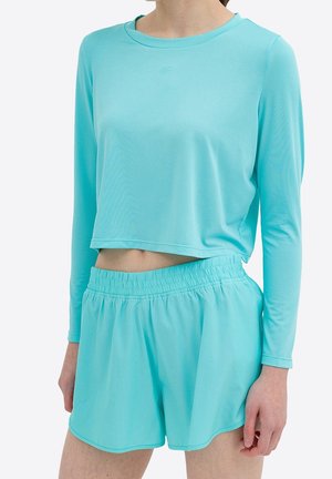 Woman wearing light blue long-sleeve cropped top and matching elastic waist shorts, standing against a plain background.