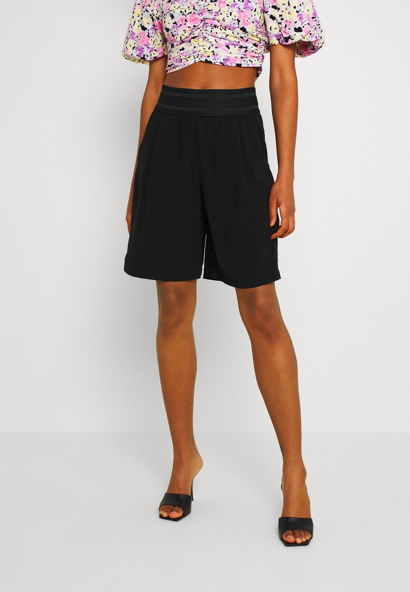 Schwarze knielange Shorts mit lässiger Passform, elastischem Bund und glatter Stoffqualität. Tragen kombiniert mit einem floral gemusterten Cropped-Top und schwarzen Sandalen.