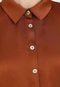 Braune Satinbluse mit Kragen, ausgestattet mit fünf runden hellcremefarbenen Knöpfen mit sichtbaren Nähten und einer glatten Textur.