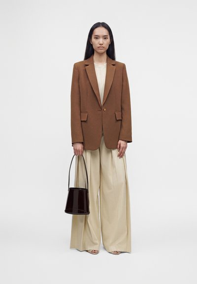 Femme debout portant un blazer marron, un pantalon large crème, un haut beige, tenant un sac seau en cuir verni noir avec de longues anses.