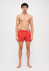 Shorts de bain rouges en tissu léger avec taille élastique. Présente un logo sur le devant et une finition lisse, sans texture.
