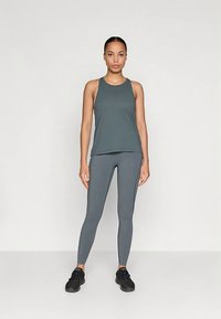 Γυναίκα που φοράει ταιριαστό γκρίζο αμάνικο μπλουζάκι και κολάν, όρθια με μαύρα αθλητικά σε απλό ανοιχτό φόντο.