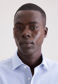 Camicia a righe con bottoni in azzurro chiaro e bianco, caratterizzata da un colletto aperto, tessuto liscio e una sottile lucentezza. Il volto del modello è al centro.