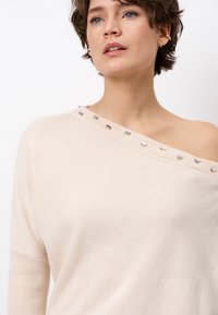 Lichtbeige off-shoulder trui van zachte stof, met metallic accenten rond de halslijn en een relaxte pasvorm. Glad textuur.