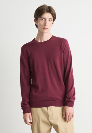 Pull-over long à manches en bordeaux en tissu doux avec col rond. Présente des poignets et un ourlet côtelés, associé à un pantalon beige clair.
