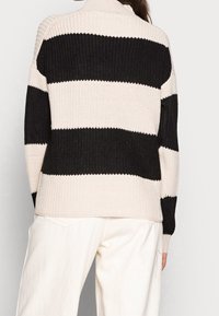 Gestrickter Pullover mit horizontalen schwarzen und cremefarbenen Streifen, hohem Kragen und geripptem Muster. Kombiniert mit cremefarbenen Hosen in lockerer Passform.