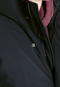 Chaqueta negra con un alto cuello, cierre de cremallera y detalle metálico a presión. Acompañada de una prenda interior color burdeos asomando por la parte del cuello.