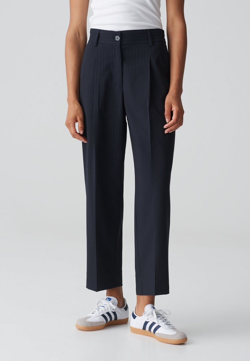 Personne portant un pantalon sur mesure à fines rayures bleu marine, un débardeur blanc et des baskets blanches avec des rayures bleu marine, debout devant un fond uni.