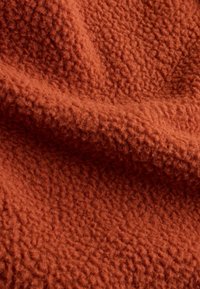 Teksturirana flis tkanina v opečnato oranžni barvi z mehko, puhasto površino. Izstopajoč, nepravilni vzorec ustvarja globino in vizualno zanimivost.