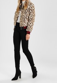 Osoba v bomber jacketu s leopardím potiskem, bílém roláku, černých skinny kalhotách a černých lodičkách na kotníky se zlatými knoflíky.