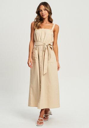 Beige sleeveless dress with a tied waist, buttons down the front, and a flowing skirt. Smooth cotton fabric, square neckline, and pockets.