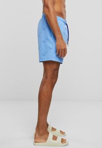 Shorts de bain pour hommes en tissu bleu clair, avec un motif bleu foncé répété. Ceinture élastique avec cordon de serrage. Sandales à glissière en vert pâle.